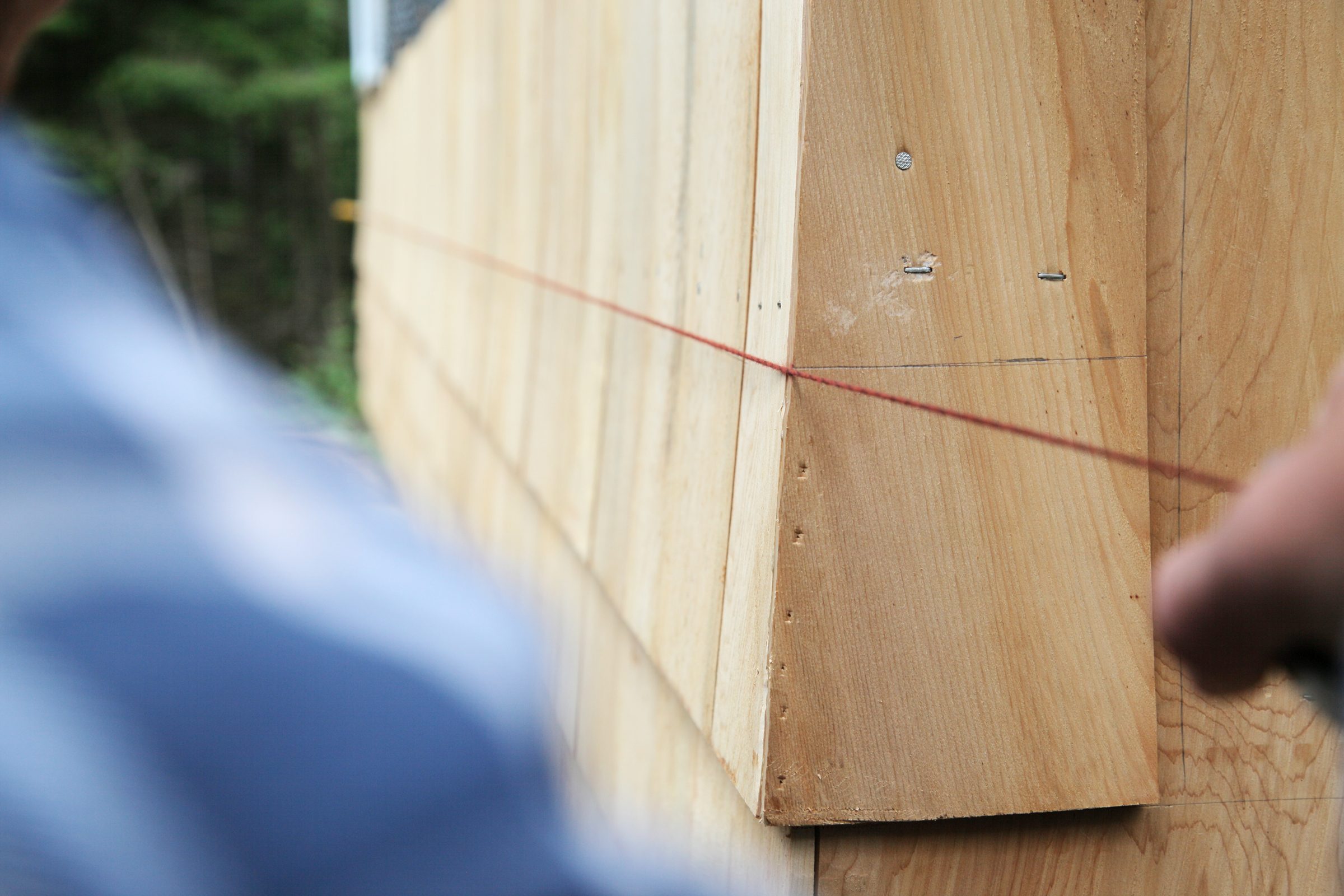 A person pulls a red string taut along wooden panels, measuring for accuracy in a construction setting surrounded by greenery.