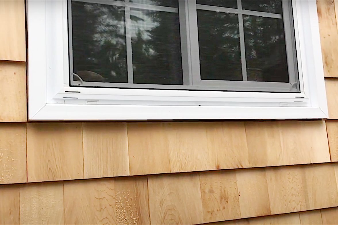 A window with a white frame is positioned above wooden shingles. The window is partially closed, surrounded by a lightly stained wooden exterior.