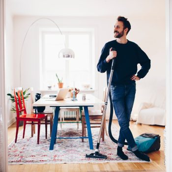 A man stands with a vacuum cleaner, smiling in a bright room. A dining table with a laptop sits nearby, surrounded by chairs and bookshelves.