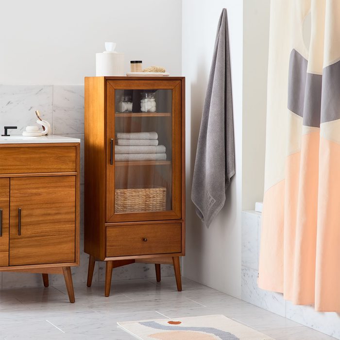 A wooden cabinet displays towels and a basket. A grey towel hangs nearby, and a colorful shower curtain adds visual interest in a bathroom setting.