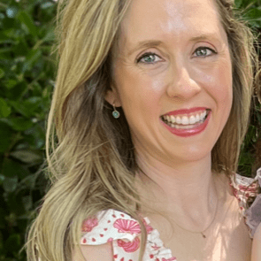 A woman with long, wavy hair smiles brightly while wearing a floral dress. Green foliage serves as the backdrop, creating a cheerful outdoor atmosphere.