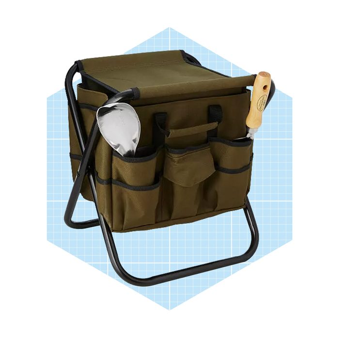 A green gardening bag with compartments holds a silver trowel and a wooden-handled tool, supported by a black frame against a light blue background.