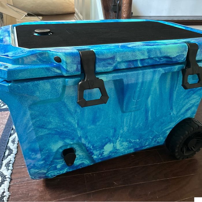 A blue, marbled cooler with black latches and wheels sits on a wooden floor, ready for use, showcasing a durable design for outdoor activities.