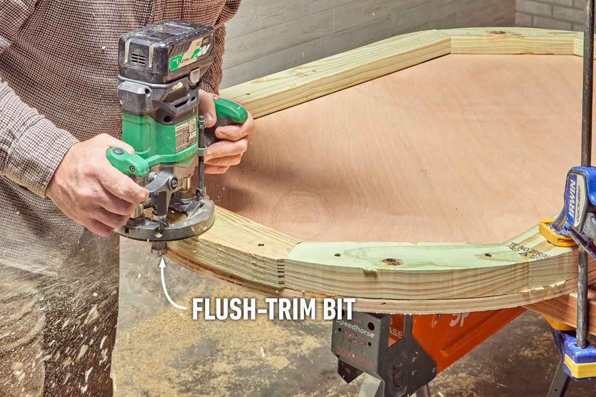 A person uses a router with a flush-trim bit to smooth the edges of wood mounted on a workbench, surrounded by sawdust and tools.