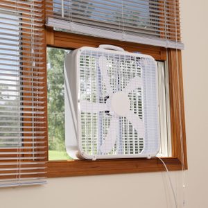 A box fan is positioned in a window, drawing air from outside, surrounded by wooden frames and blinds, with green foliage visible beyond the glass.