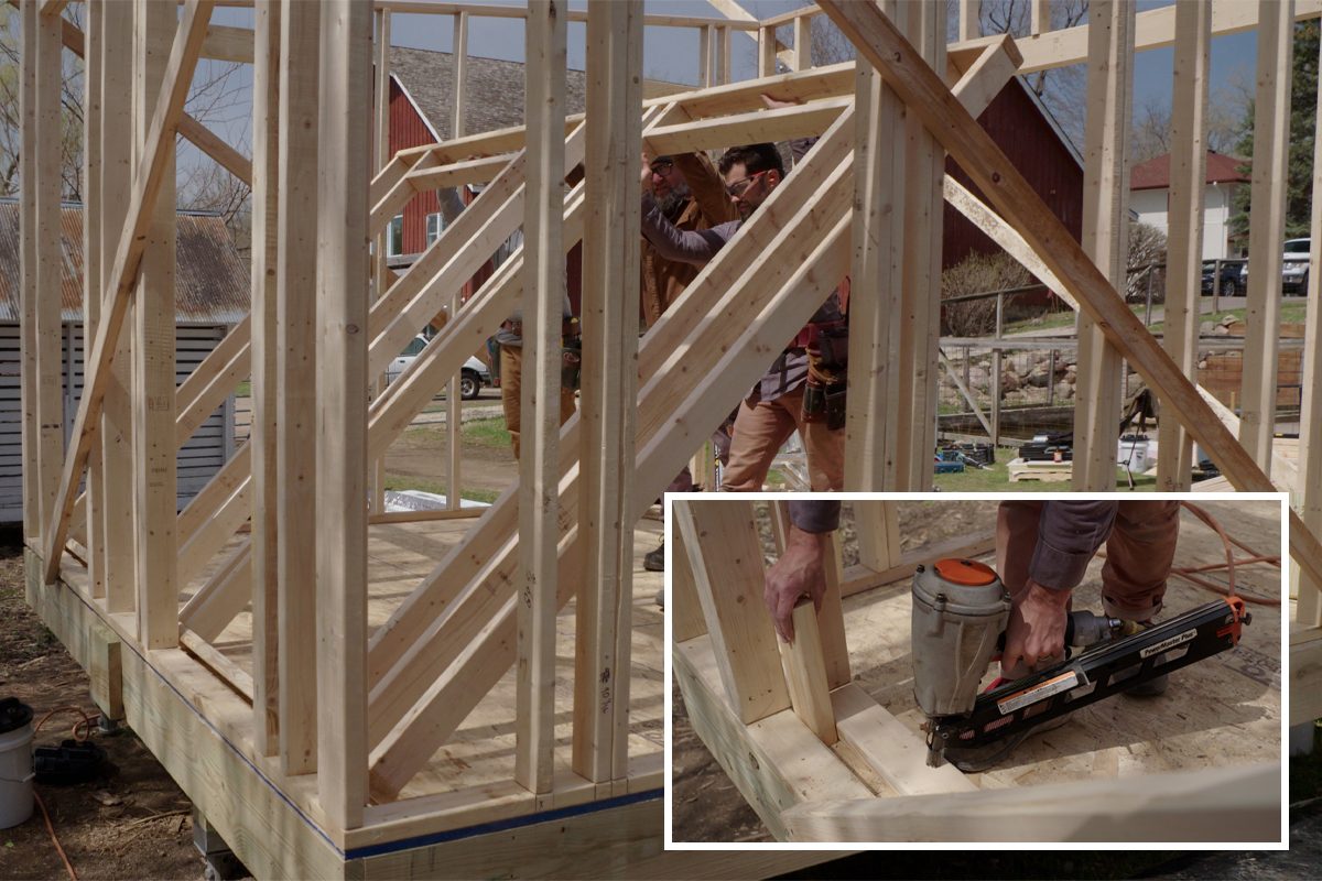 Framed wooden structure is being assembled. A worker uses a nail gun to secure beams. Surrounding area includes tools and nearby buildings in daylight.