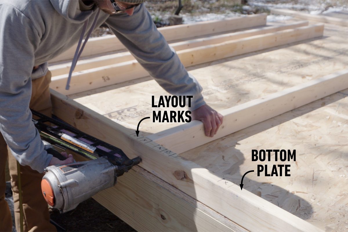 A person uses a nail gun to secure a wooden bottom plate, aligning it with marked layout points on a construction platform in a sunny outdoor setting.