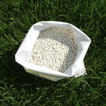 A bag filled with small white pellets rests on green grass, suggesting use in gardening or fertilization. The bag is partially open at the top.