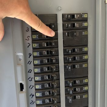 A hand points at a circuit breaker panel, with various switches in the "ON" and "OFF" positions, mounted on a metallic surface.
