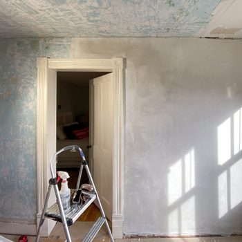 A step ladder stands in a partially painted room, with a doorway leading to another space and sunlight casting shadows on the wall.