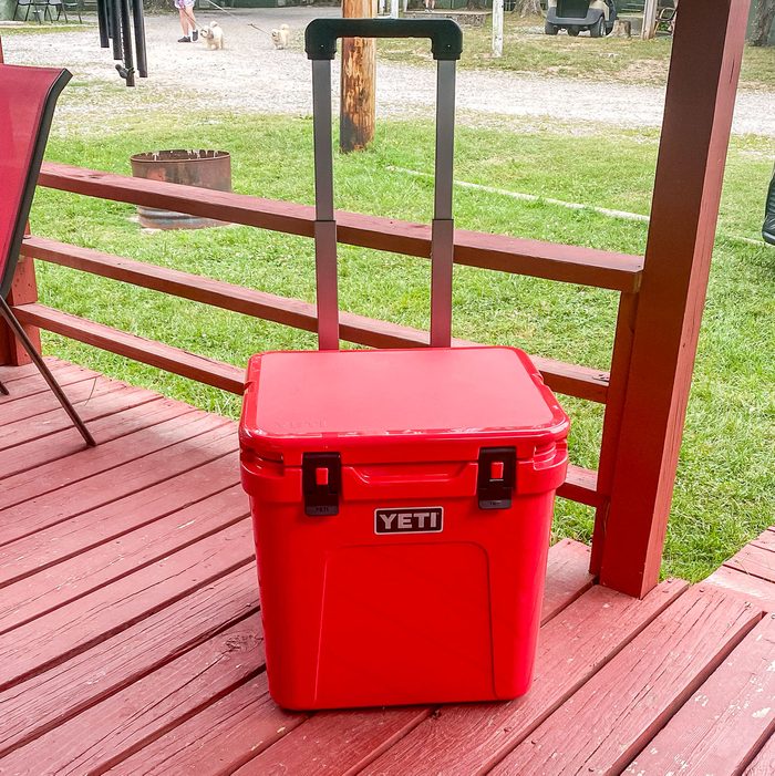 A red YETI cooler stands on a wooden porch with an extended handle, while green grass and a gravel path are visible in the background.