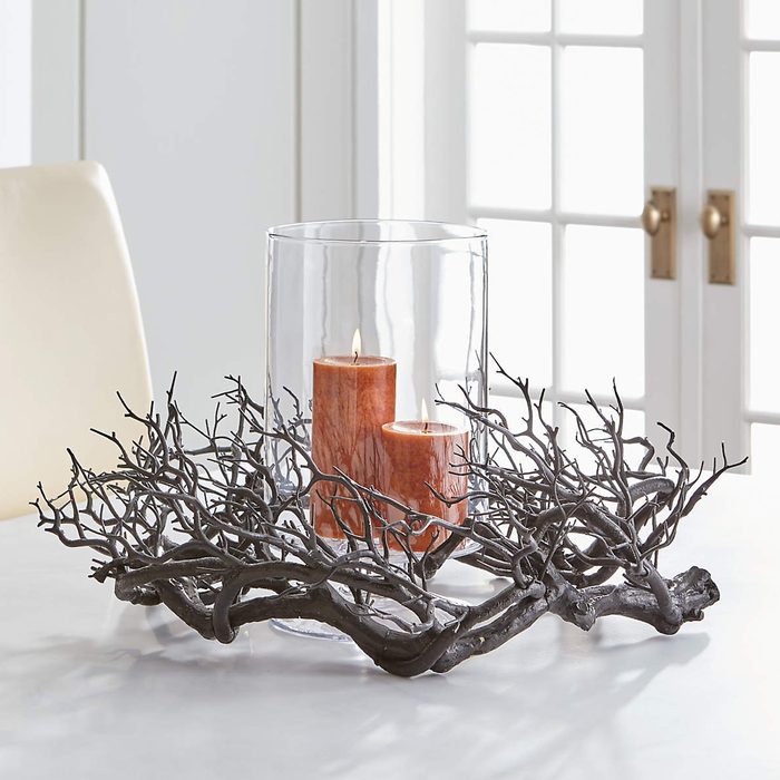 A decorative candle holder features two orange candles inside a glass cylinder, surrounded by an artistic, dark, branch-like base on a table.
