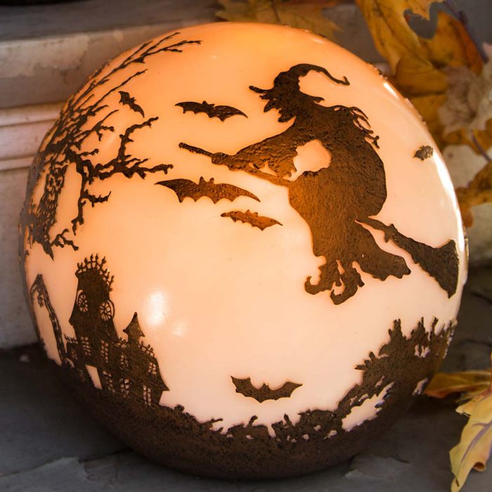 A round, illuminated Halloween decoration depicts a witch on a broomstick, surrounded by bats and a haunted house, set against a soft orange background.