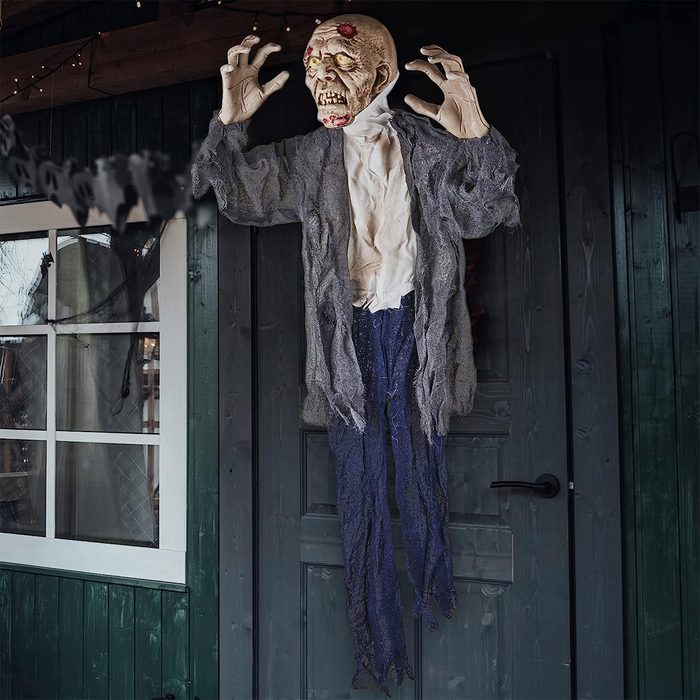 A zombie figure hangs on a door, arms outstretched, with a terrifying expression, set against a rustic wooden backdrop with a window nearby.
