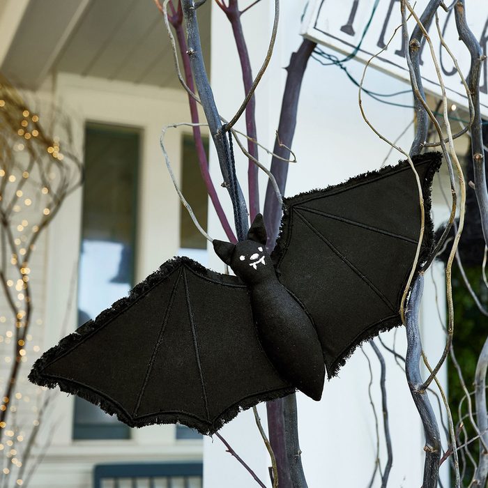A large, black, plush bat hangs from slender, dark branches, decorated with soft textures, in a bright outdoor setting near a home.