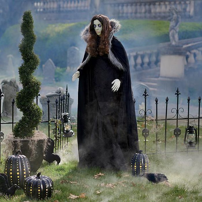 A lifelike figure in a black cloak stands amidst fog, near decorative pumpkins and a spiraled shrub, in a graveyard setting with tombstones and a fence.