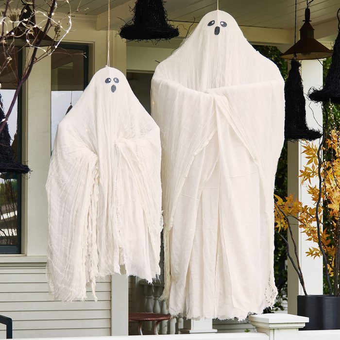 Two white ghost decorations hang from a porch ceiling, showcasing simple black eyes and mouths, surrounded by dark hanging decorations and autumn foliage.