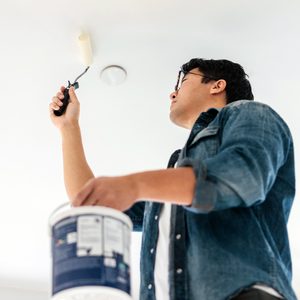 A person in a denim shirt and glasses is painting a ceiling with a roller. They hold a paint can in one hand and reach upward. The perspective is from below, looking up at the person and the ceiling.